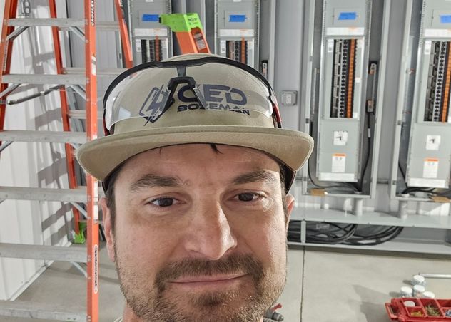 Electrician wearing hard hat and safety harness in front of newly installed commercial service panels at a Bozeman job site